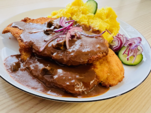 panierte hähnchenschnitel auf einem weißen teller, bedeckt mit brauner glänzender rahmsauce, sowie kartoffelbrei