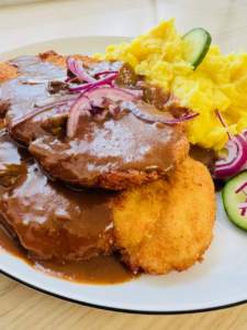 panierte hähnchenschnitel auf einem weißen teller, bedeckt mit brauner glänzender rahmsauce, sowie kartoffelbrei