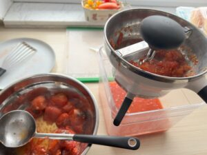 schüssel mit schöpfkelle und gerösteten tomaten, daneben großer behälter mit passiermühle oben drauf